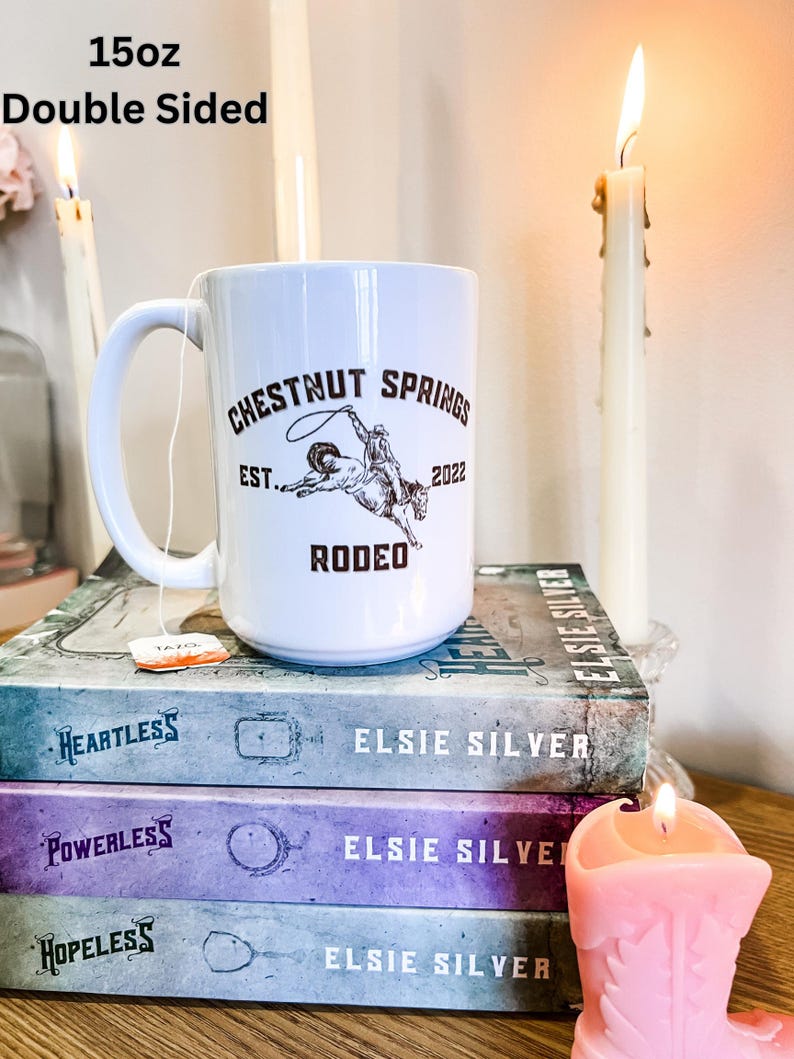 May include: White ceramic mug with a brown graphic of a cowboy riding a bucking bronco. The graphic includes the text "Chestnut Springs Est. 2022 Rodeo". The mug is sitting on a stack of three books with the title "Elsie Silver" on each book. There is a pink cowboy boot candle next to the mug.
