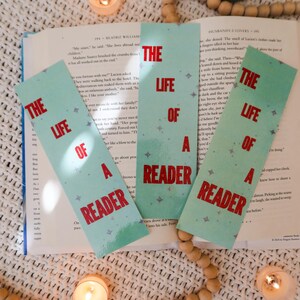 Puede incluir: Tres marcadores turquesa con el texto "THE LIFE OF A READER" en rojo, sobre un libro abierto. La escena incluye velas encendidas y cuentas de madera, creando un ambiente de lectura acogedor.