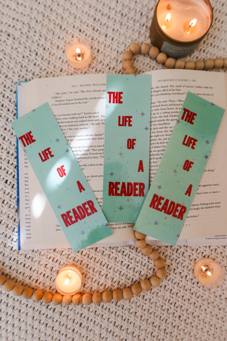 Puede incluir: Tres marcadores turquesa con el texto "THE LIFE OF A READER" en rojo, sobre un libro abierto. La escena incluye velas encendidas y cuentas de madera, creando un ambiente de lectura acogedor.