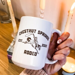 May include: White ceramic mug with a brown graphic of a cowboy riding a horse. The graphic includes the text "Chestnut Springs Est. 2022 Rodeo". The mug is 15 oz and double-sided.