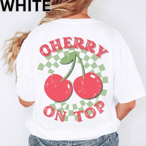 May include: White t-shirt with a red and green graphic of two cherries with stems and leaves. The graphic is surrounded by a checkered pattern and the text "CHERRY ON TOP" is below the cherries.