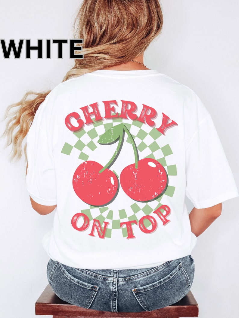 May include: White t-shirt with a red and green graphic of two cherries with stems and leaves. The graphic is surrounded by a checkered pattern and the text "CHERRY ON TOP" is below the cherries.