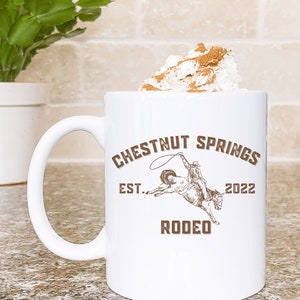 May include: White ceramic mug with a brown graphic of a cowboy riding a horse. The graphic says "Chestnut Springs Est. 2022 Rodeo". The mug is 11 ounces and double-sided.