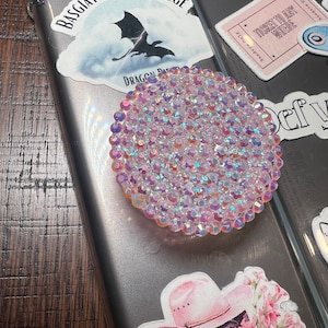 May include: A round, iridescent crystal phone grip is affixed to a phone case. The grip is surrounded by stickers, including one with a dragon and the text "BASGIATH WAR COLLEGE". Other stickers include text that reads "Hello, Feyre D" and "TICKET TO ANY WHERE".