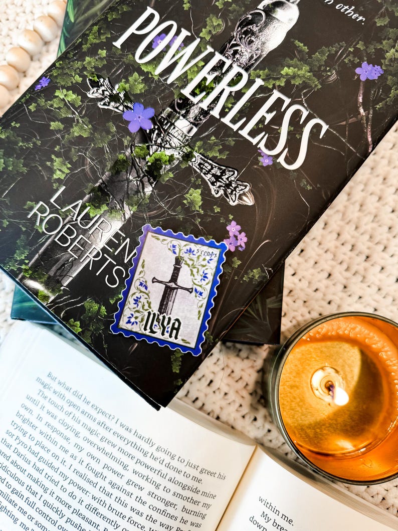May include: A close-up of a book titled "Powerless" by Lauren Roberts. The book cover features a sword with a black background and purple flowers. A sticker with a sword and the word "Ilya" is on the book.
