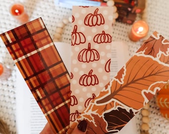 May include: Three decorative bookmarks with fall-themed designs. One features a plaid pattern in shades of orange and brown. Another has a pumpkin pattern on a pink background. The third has an autumn leaf design.