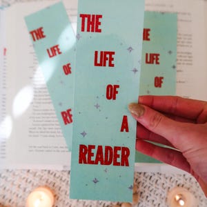 Puede incluir: Tres marcap&aacute;ginas azul claro con el texto "THE LIFE OF A READER" en rojo. Los marcap&aacute;ginas tienen un patr&oacute;n de estrellas y se sostienen cerca de un libro abierto, con velas y cuentas de madera en el fondo.