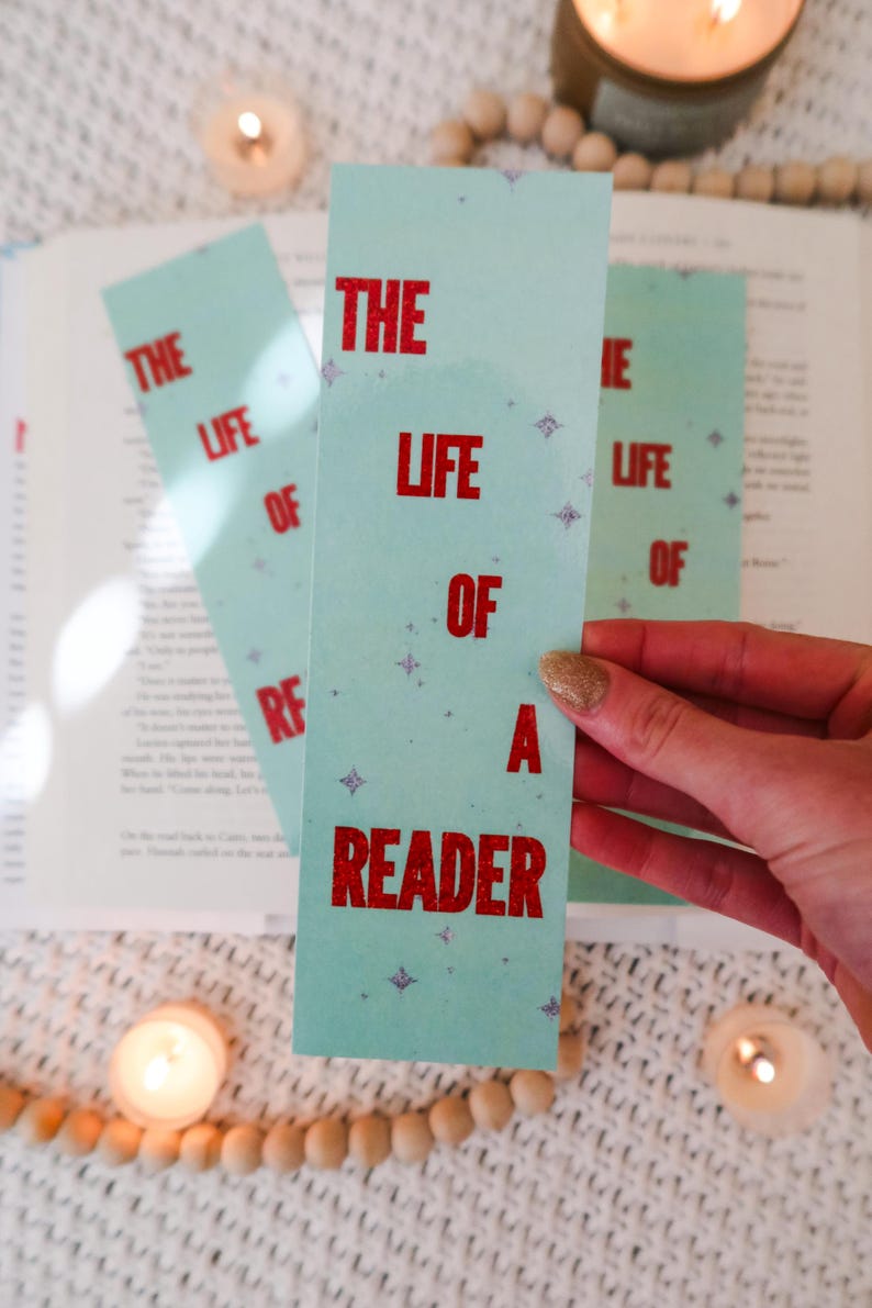 Puede incluir: Tres marcap&aacute;ginas azul claro con el texto "THE LIFE OF A READER" en rojo. Los marcap&aacute;ginas tienen un patr&oacute;n de estrellas y se sostienen cerca de un libro abierto, con velas y cuentas de madera en el fondo.