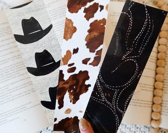 Puede incluir: Tres marcapáginas rectangulares con diseños del oeste. Uno presenta sombreros de vaquero negros sobre un fondo texturizado, otro tiene un patrón de piel de vaca y el tercero un diseño de cuero negro con costuras blancas. Se sostienen sobre un libro abierto.