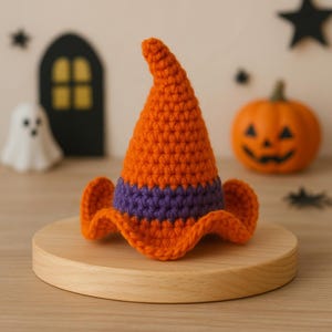 Op de afbeelding: Een gehaakte oranje heksenhoed met een paarse band en een golvende rand. De hoed wordt getoond op een houten standaard, met een uitgesneden pompoen en andere Halloween-decoraties op de achtergrond.