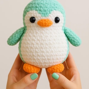 Può includere: Un giocattolo a forma di pinguino fatto a mano all'uncinetto. Il pinguino ha testa e ali turchesi, corpo bianco e piedi e becco arancioni. Ha occhi di sicurezza neri. Il giocattolo è tenuto tra le mani di una persona.