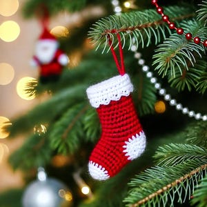 Puede incluir: Un adorno de calcetín de Navidad de crochet rojo y blanco colgado de un árbol de Navidad. El calcetín tiene un borde de crochet blanco y un diseño de copo de nieve blanco.