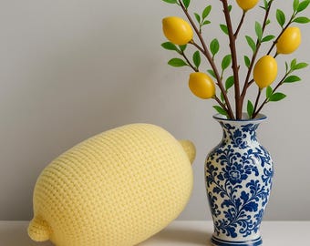 May include: A yellow crocheted lemon-shaped pillow rests beside a blue and white floral vase holding a branch with yellow lemon decorations and green leaves. The pillow is a soft, textured design.