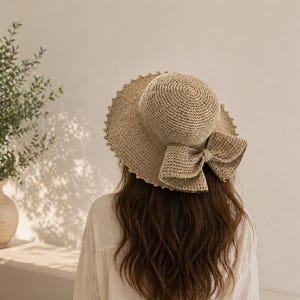 Puede incluir: Un sombrero de sol beige tejido con ala ancha y un lazo grande en el lateral. El sombrero tiene un borde festoneado y está hecho de fibra natural. El sombrero lo lleva una persona con pelo largo castaño.