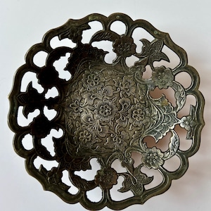 May include: Ornate, antique silver-colored metal dish with a floral pattern. The center of the dish features a detailed floral design, while the rim has a cut-out floral and leaf design. The dish is round and has a slightly raised edge.