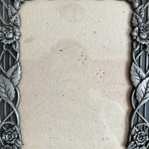 May include: A rectangular, ornate picture frame with a floral design. The frame is made of a dark, possibly metal material, with roses and leaves adorning the edges. The center is empty, ready for a photo.