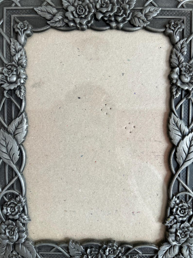 May include: A rectangular, ornate picture frame with a floral design. The frame is made of a dark, possibly metal material, with roses and leaves adorning the edges. The center is empty, ready for a photo.