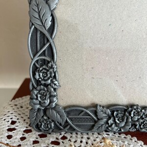 May include: A decorative silver-colored picture frame with a floral and leaf design. The frame features detailed roses and leaves, with an intricate pattern along the edges. The frame is designed to hold a photograph or artwork.