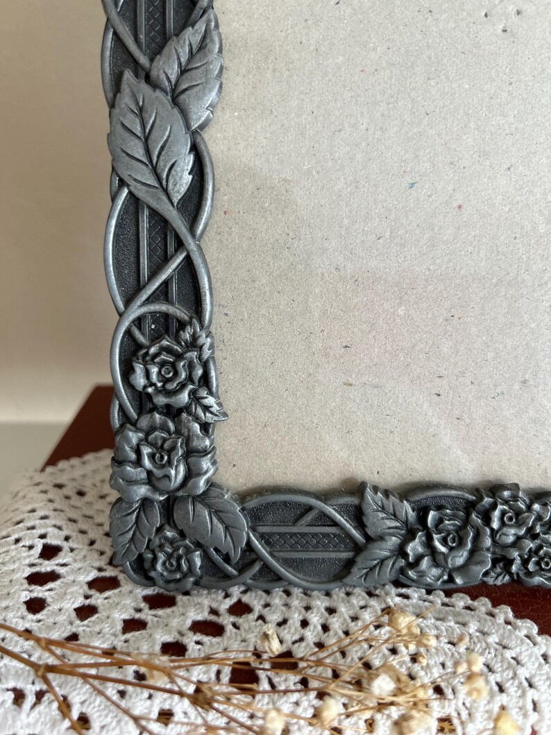May include: A decorative silver-colored picture frame with a floral and leaf design. The frame features detailed roses and leaves, with an intricate pattern along the edges. The frame is designed to hold a photograph or artwork.