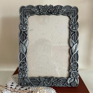 May include: A rectangular, ornate silver-toned picture frame. The frame is decorated with roses and leaves, with a braided design along the edges. The frame is empty, ready for a photo. The frame is sitting on a red book.