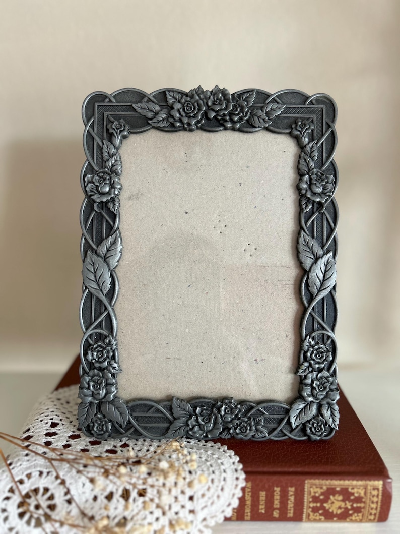 May include: A rectangular, ornate silver-toned picture frame. The frame is decorated with roses and leaves, with a braided design along the edges. The frame is empty, ready for a photo. The frame is sitting on a red book.