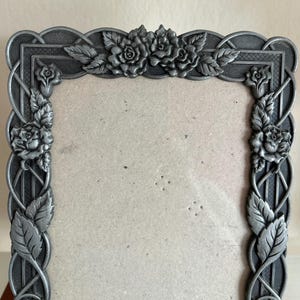May include: A decorative metal picture frame with a gray finish. The frame features a floral design with roses and leaves. The frame has an ornate, vintage style and is designed to hold a photograph or artwork.