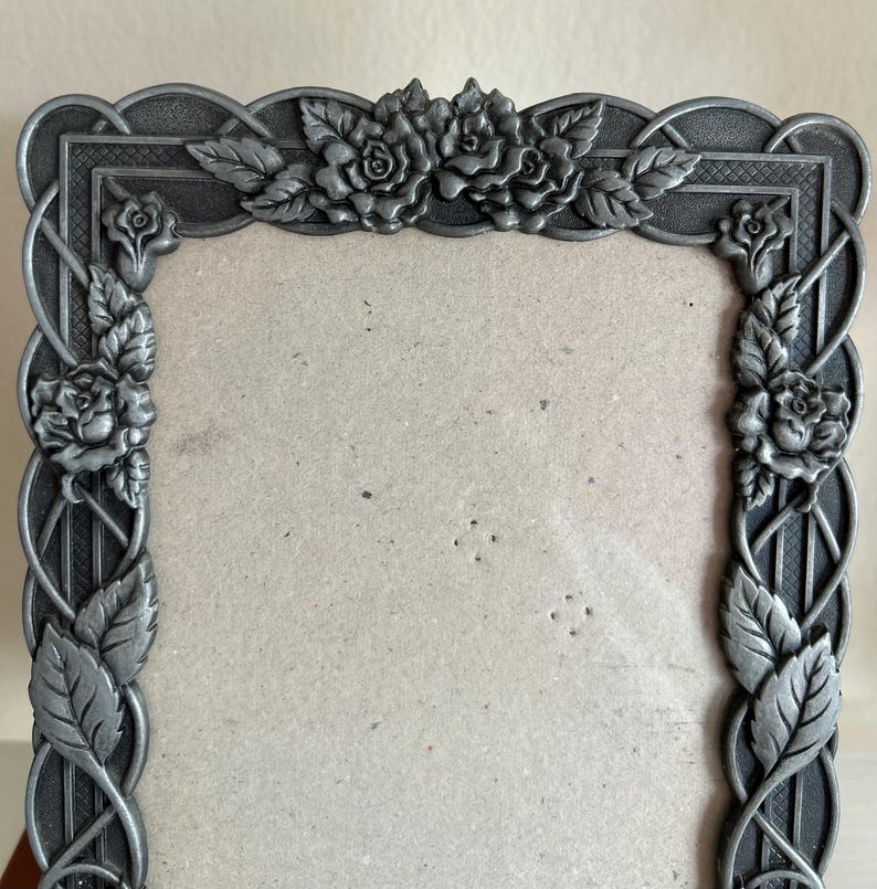 May include: A decorative metal picture frame with a gray finish. The frame features a floral design with roses and leaves. The frame has an ornate, vintage style and is designed to hold a photograph or artwork.