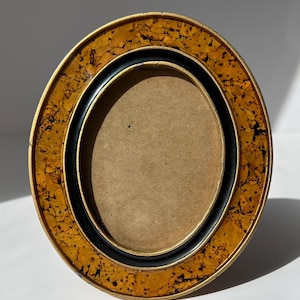 May include: Oval picture frame with a gold and black design. The frame has a textured, speckled gold outer ring, a black inner ring, and a brown backing. The frame is designed to hold an oval-shaped photograph.