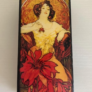 May include: A wooden box with a colorful image of a woman in a yellow dress and red flowers. The image is in the Art Nouveau style and features a geometric pattern in the background. The box is signed "Mucha" in the lower right corner.