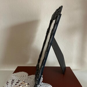 May include: A dark blue, ornate picture frame with a decorative border, standing on a red book. The frame is propped up by a stand on the back. A white lace doily and dried flowers are in the foreground.