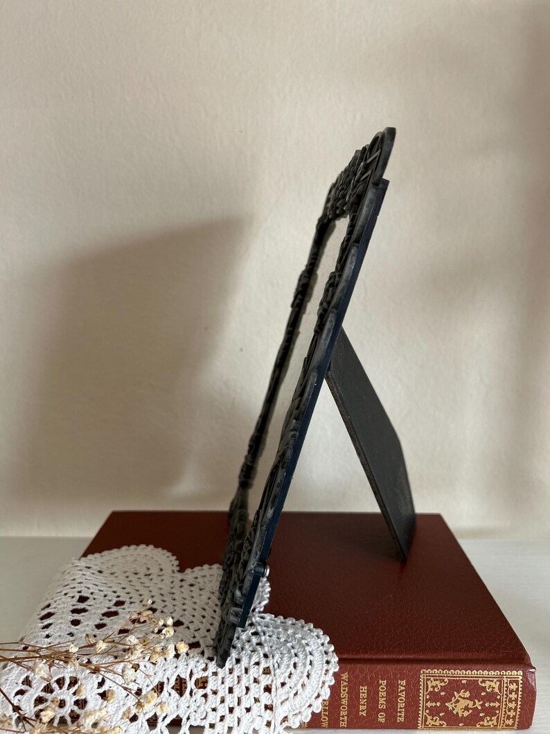 May include: A dark blue, ornate picture frame with a decorative border, standing on a red book. The frame is propped up by a stand on the back. A white lace doily and dried flowers are in the foreground.