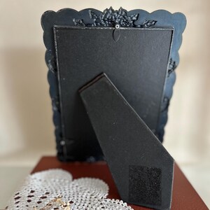 May include: A black, ornate picture frame with a scalloped edge and decorative floral accents. The back of the frame is visible, showing a black easel stand and a black backing. The frame is resting on a red book and a white doily.