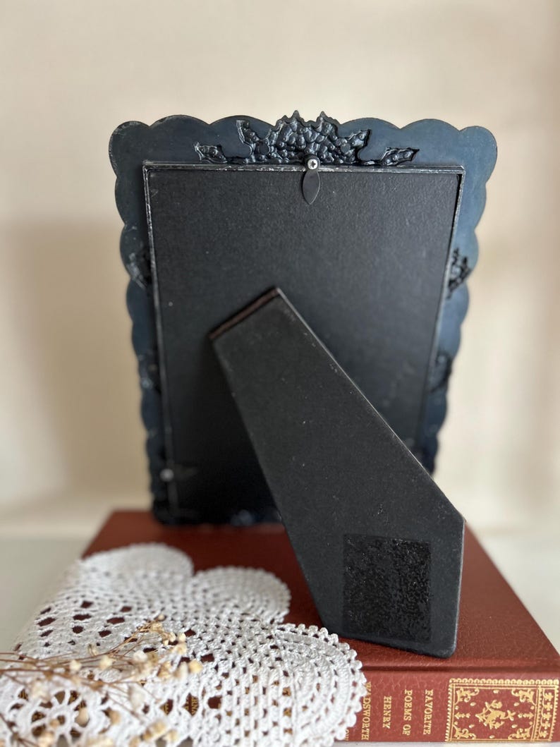 May include: A black, ornate picture frame with a scalloped edge and decorative floral accents. The back of the frame is visible, showing a black easel stand and a black backing. The frame is resting on a red book and a white doily.