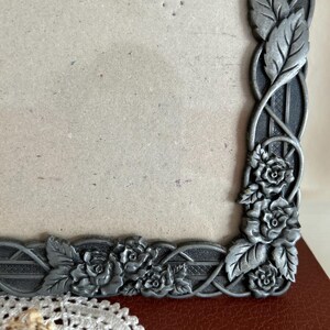 May include: A detailed close-up of a decorative metal picture frame. The frame is silver-toned with an intricate design of roses and leaves. The frame is resting on a red surface.