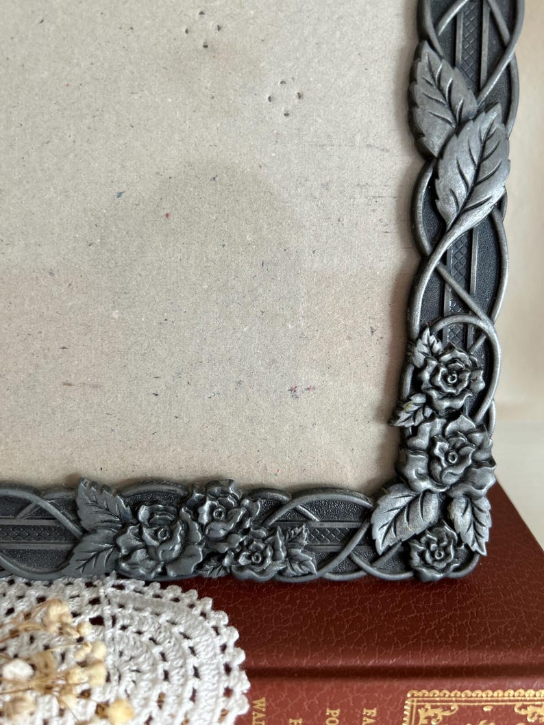 May include: A detailed close-up of a decorative metal picture frame. The frame is silver-toned with an intricate design of roses and leaves. The frame is resting on a red surface.