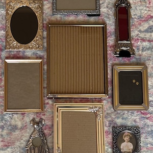 May include: A collection of various antique picture frames in gold, silver, and bronze tones. The frames are in different shapes and sizes, including oval, rectangular, and a dress-shaped frame. Some frames have ornate detailing.