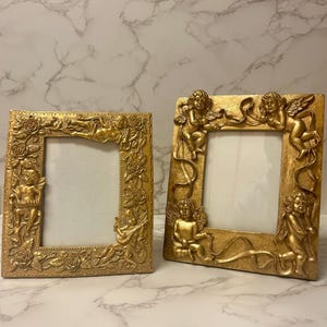 May include: Two gold picture frames with ornate cherub and floral designs. The frames are both rectangular and have a raised edge around the opening for the picture.