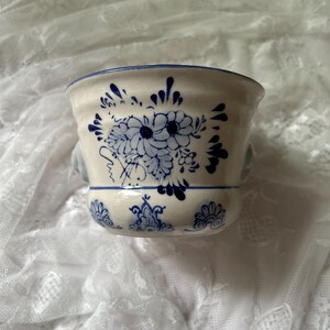 May include: A cream-colored ceramic bowl featuring a hand-painted blue floral pattern and decorative motifs. The bowl has a wide, flared rim with a thin blue line along the edge and a rounded base, showcasing a classic delft-style aesthetic.