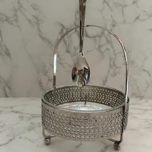 May include: A silver-colored spoon rests in a matching holder. The holder has a decorative, openwork design around a clear glass bowl. The spoon is suspended from a curved handle, creating an elegant display.
