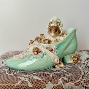 May include: A ceramic figurine of a shoe in a light teal color, adorned with a pink bow and lace details. The shoe is filled with several small, brown mice. One mouse is wearing a bonnet. A charming decorative piece.