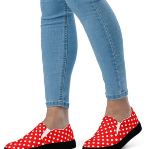 May include: Red slip-on shoes with white polka dots and black soles. The shoes are worn with light blue denim jeans. The shoes have a classic, casual style.