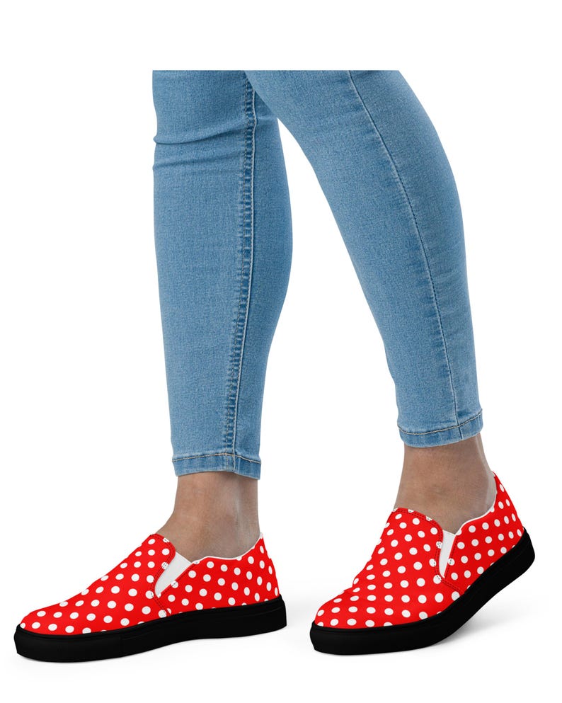 May include: Red slip-on shoes with white polka dots and black soles. The shoes are worn with light blue denim jeans. The shoes have a classic, casual style.