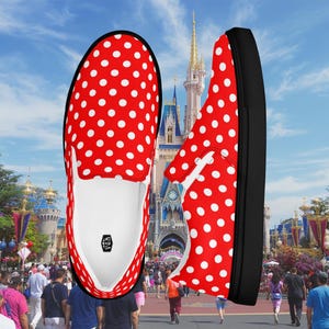 May include: Red and white polka dot slip-on shoes with black soles. The shoes are displayed against a background of a theme park with a castle and people. The shoes have a white interior and a logo.