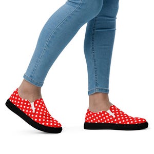 May include: Red slip-on sneakers with white polka dots and black soles. The shoes are worn with light blue denim jeans. The sneakers have a classic, casual style.