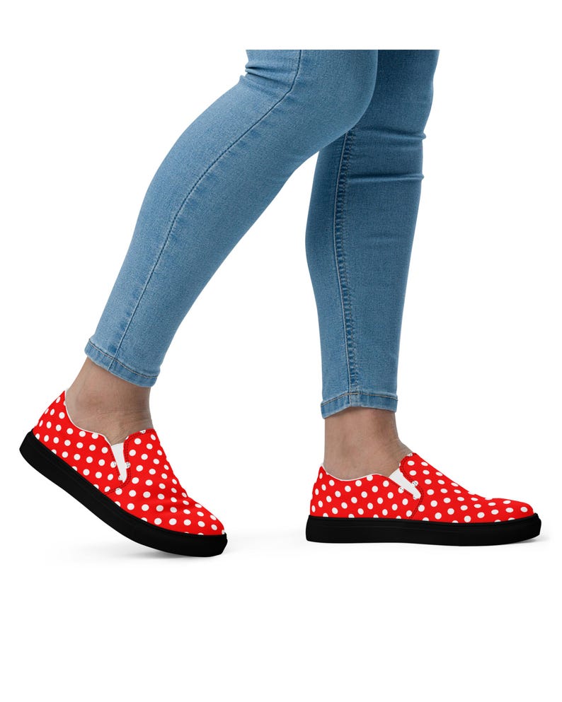 May include: Red slip-on sneakers with white polka dots and black soles. The shoes are worn with light blue denim jeans. The sneakers have a classic, casual style.