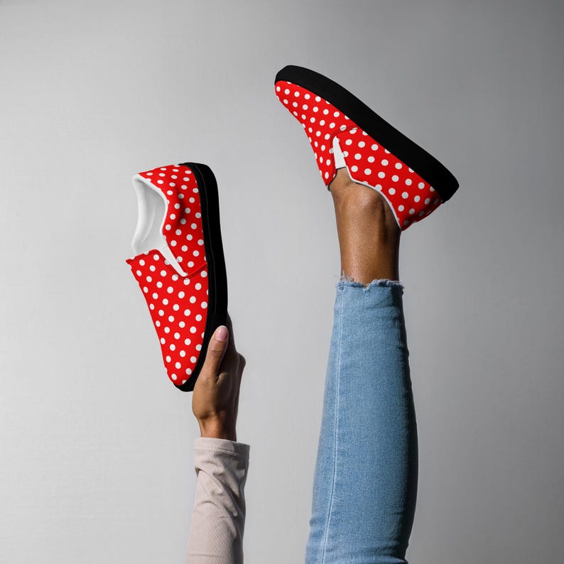 May include: Red slip-on shoes with white polka dots and black soles. The shoes have a classic design and are made of a durable material. The interior is white. The shoes are held up in the air.