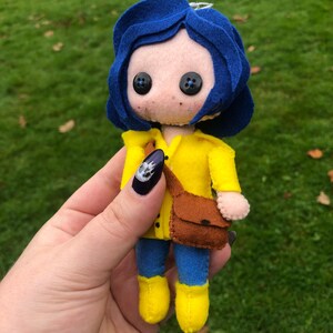 May include: A felt doll of a girl with blue hair, wearing a yellow raincoat, blue jeans, and yellow boots. She is carrying a brown bag.