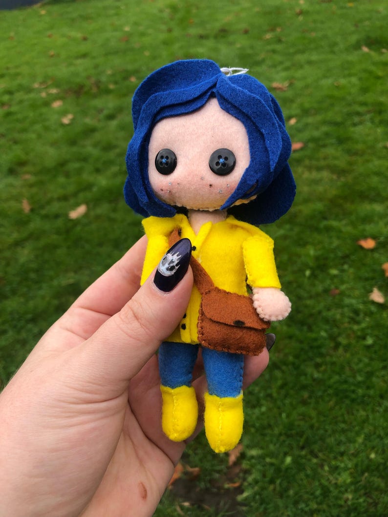 May include: A felt doll of a girl with blue hair, wearing a yellow raincoat, blue jeans, and yellow boots. She is carrying a brown bag.