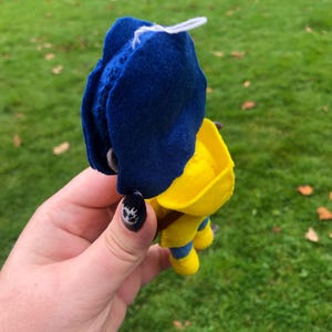 May include: A felt toy with a blue hat and yellow body. The toy is wearing a yellow raincoat and has a white string attached to the top of its head.