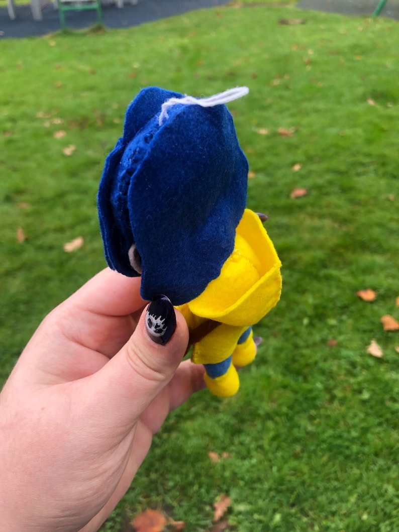 May include: A felt toy with a blue hat and yellow body. The toy is wearing a yellow raincoat and has a white string attached to the top of its head.
